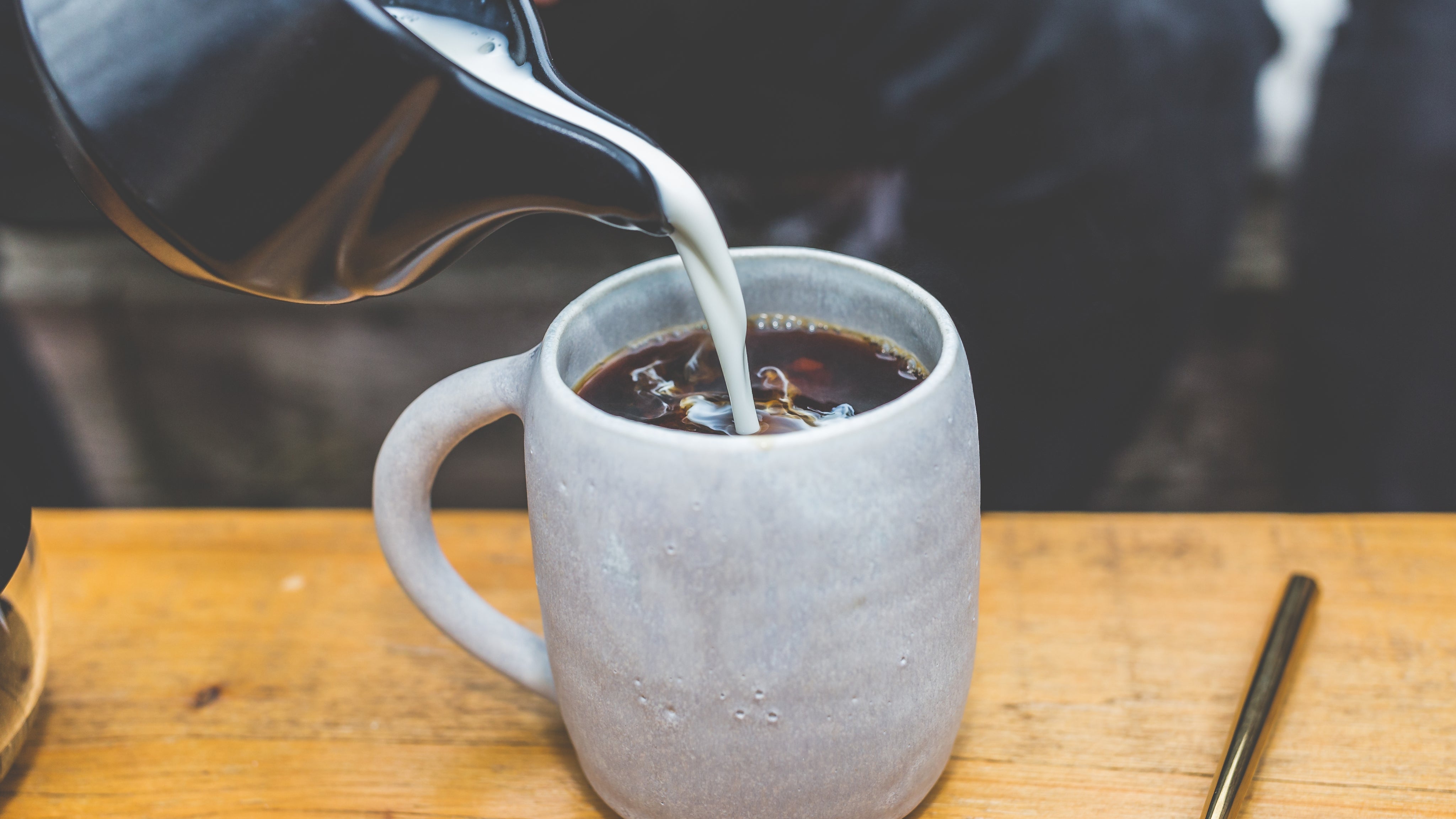 Person pouring coffee from a pot into a gray mug on a wooden table.