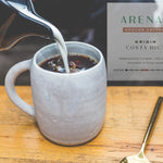 Coffee being poured into a mug with Arenal Costa Rica coffee packaging in the background.